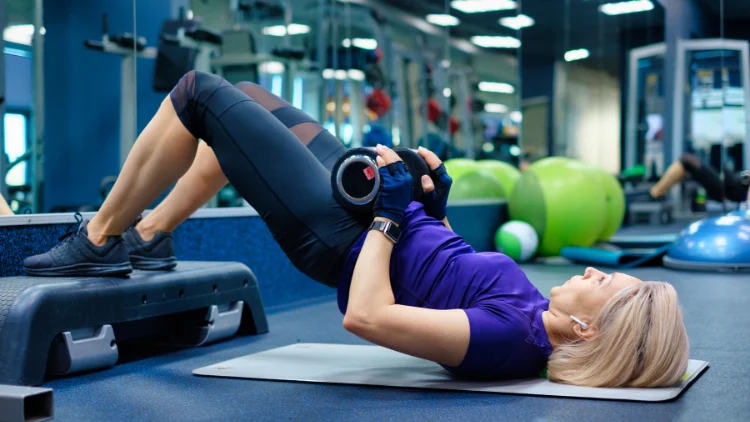 A blonde woman wearing workout clothes is lying on a white exercise mat, she has a dumbbell resting on her hips and is lifting her hips up towards the ceiling in a hip thrust motion.