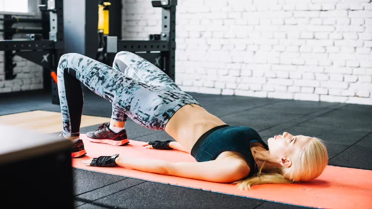 Blonde woman lying on a orange mat in black top and pattern leggings lifting her waist in the gym.