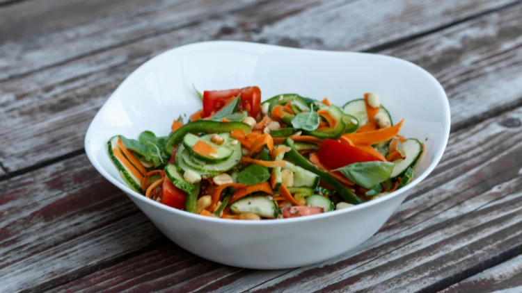 On a white tabletop is a dish of crunchy Thai peanut salad that has been made with a colorful mix of tomato, green bell peppers, carrots, and cucumber.