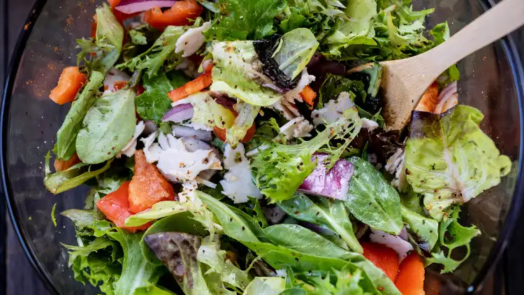 A bowl of colorful healthy salad containing mixed greens, cherry tomatoes, cucumbers, carrots, and red onions, served with a wooden spoon on the side.