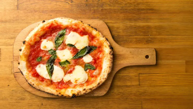 A round, baked pizza is placed on a wooden board lying on a wooden table, with tomato sauce, mozzarella cheese, and basil on top.