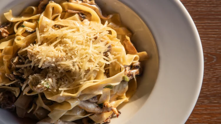 Satisfying fettuccine noodles coated in a creamy cheese sauce, topped with pieces of chicken, garnished with grated cheese, served on a white plate placed on a wooden table-like surface.
