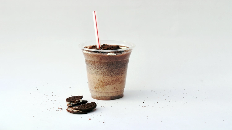 A cup filled with a creamy, chocolate-colored shake, topped with crushed cookies and chocolate shavings, and a red-and-white striped straw are visible, with chopped chocolate cookies on a white background.