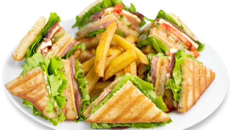 A club sandwich cut diagonally into quarters and arranged on a plate with a side of golden-brown french fries in the center, the sandwich is made with layers of turkey, ham, bacon, lettuce, tomato, and mayonnaise, stacked between three slices of toasted bread.