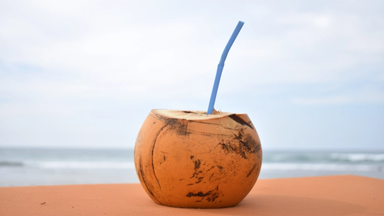 A brown coconut with a rough, fibrous exterior and a blue bent straw sitting on a sandy, beach-like surface, with a view of the ocean in the background.