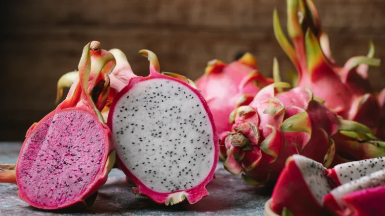 A vibrant, pink and white dragon fruit sliced in half, displaying its speckled, juicy interior, with an unsliced dragon fruit placed beside it on a brown background.