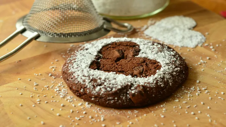 A dark chocolate chip cookie with sweetened milk powder in the shape of a heart is placed on a wooden board, with a stainless steel flour sieve sifter and white heart shape powder visible in the background.