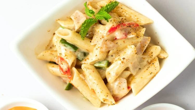 A bowl of creamy and saucy penne noodles, topped with tomatoes, green bell peppers, and fresh parsley, is placed on a white background, with two white plates in the background.