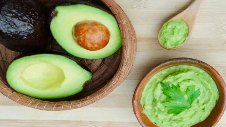 A wooden bowl and spoon filled with mashed avocado and half slice of avocado on the side.