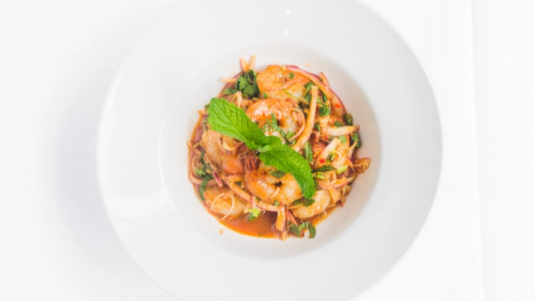 A top view photo of a serving of spicy Thai shrimp salad made with shrimp, greens, and vegetables served on a white plate.