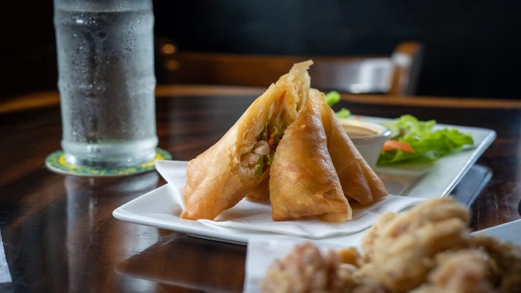On a white plate is a golden-brown pork egg roll, crispy on the outside and filled with savory pork, vegetables, and seasonings, accompanied by a small dipping sauce container on the side and a glass of cold water in the background, creating a refreshing and light pairing for the savory egg roll.