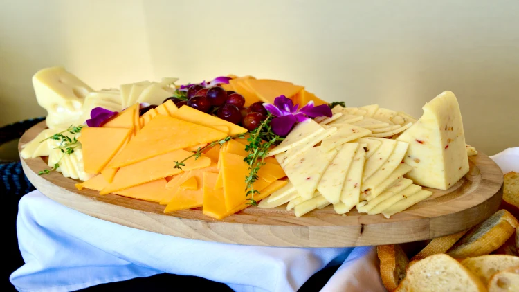 Triangular slices of cheddar cheese, gouda cheese, and emmental cheese are arranged alongside grapes and violet flowers on a wooden board with a white tablecloth and sliced bread in the background.