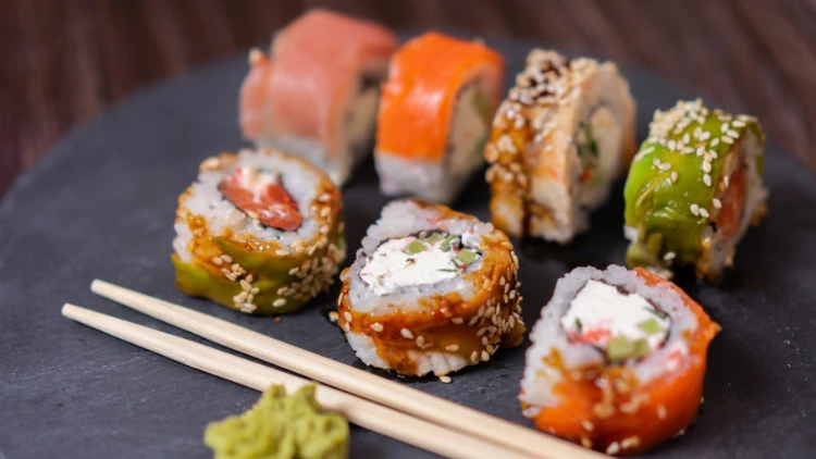 A plate of seven arranged sushi rolls with a variety of fillings and toppings, accompanied by chopsticks resting on the plate and a small, green wasabi, presents a visually appealing and flavorful meal.