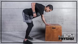 A man wearing a black shirt, gray shorts, black tights, and black shoes is performing a dumbbell row exercise while standing on a concrete floor, he has a watch on his left wrist and his right hand is holding a dumbbell while his left hand is resting on a wooden box beside him.
