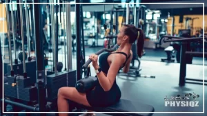 A fit woman wearing gym clothes and a watch with ponytail hair is performing a lat pulldown exercise, one of the dumbbell pullover alternatives, and there are various gym equipment that can be seen in the background.