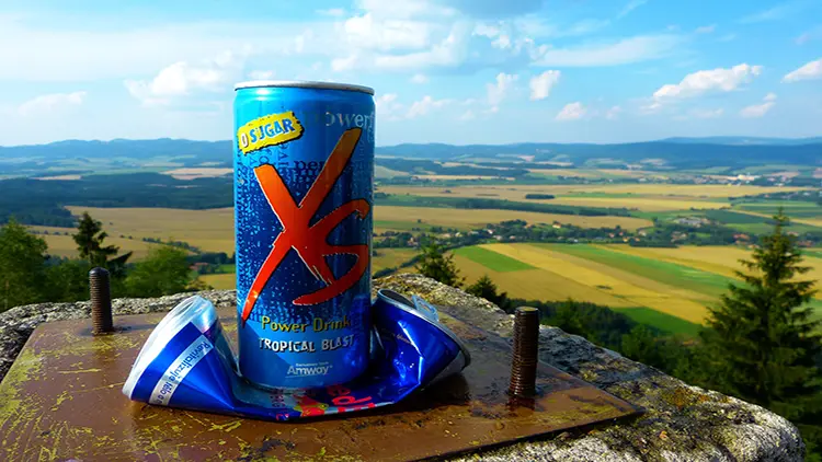 A blue can of power drink in tropical blast flavor placed on top of a flattened can of the same variety displayed on top of a concrete platform with a view of the country side, valleys, trees and the sky.