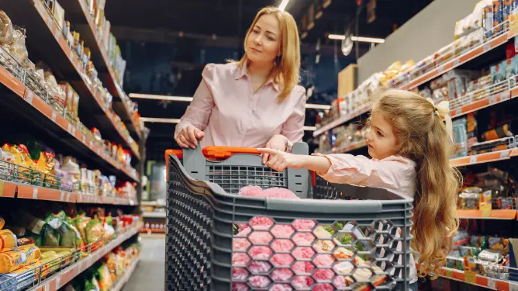 A blonde woman with a blonde girl holding on to the shopping cart while looking at the shelves with various items at a grocery store.