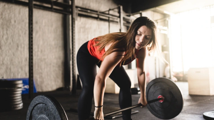 Woman in orange top and black leggings will perform deadlift exercise with plate weights in the background.