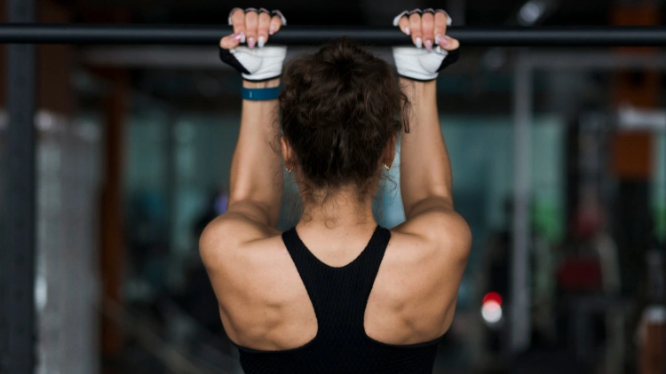 A woman in black top doing narrow pull up at the gym with white gloves in her hand and blur background of the gym.