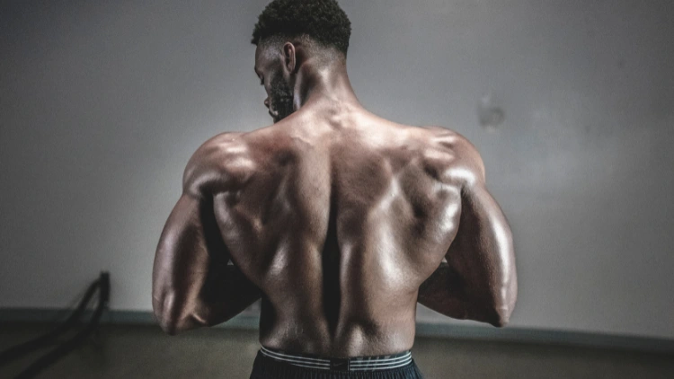 A muscular, topless man stands with his back to the camera, showcasing his impressive physique with broad, tapered shoulders, defined sections of muscle running from the base of his neck to the top of his lower back, tanned and smooth skin with veins visible beneath the surface, and powerful contractions that visibly ripple with strength, demonstrating his dedication to fitness.