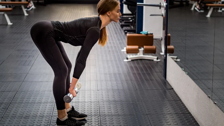 A woman wearing a black sweatshirt, black gym gym pants, and black shoes executes a dumbbell romanian deadlift in the gym, in front of her is a mirror so she can she herself while she performs the exercise.