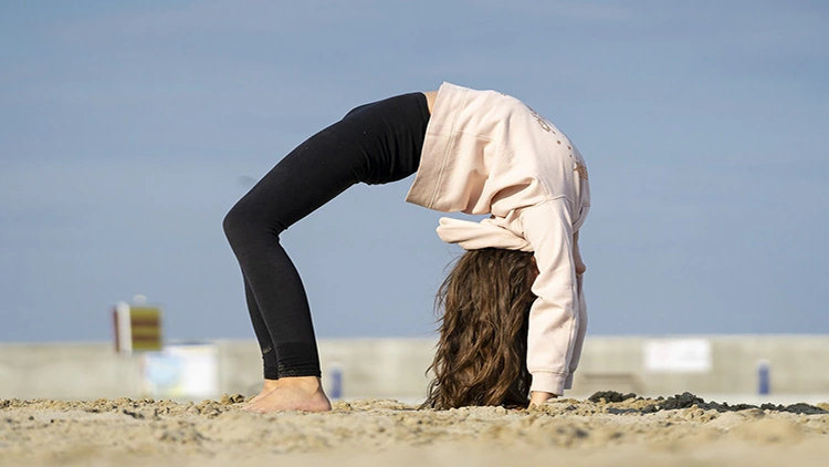 A young woman wearing a peach hoodie and black pants bending her back at the beach.