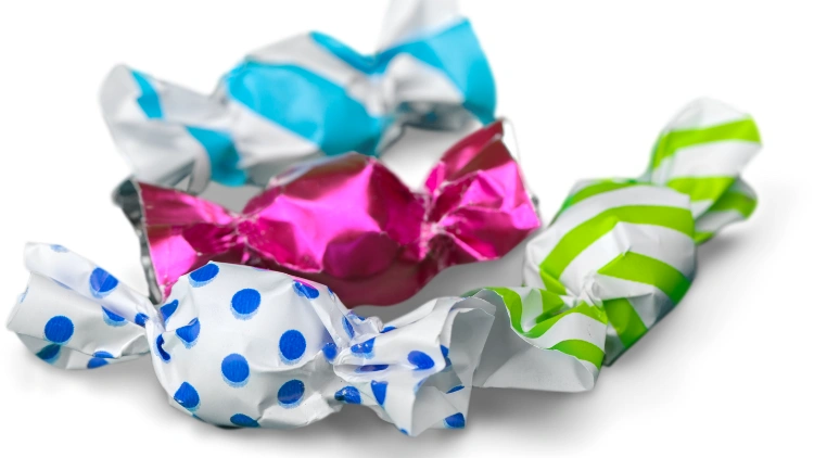 Four candies with different wrapper designs, the first candy has a solid pink wrapper, the two candy has a wrapper with stripes design, and the third candy has a wrapper with a blue polka dots pattern, the candies are arranged in a row on a flat surface.