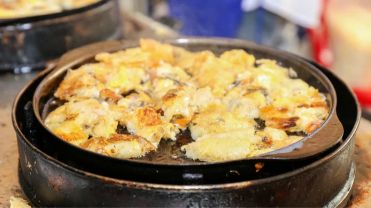 A sizzling pan of Hoi Tod, a popular Thai street food dish, with smoke rising from the pan, the dish consists of a crispy pancake made with rice flour, eggs, and a mix of seafood, usually mussels or oysters.