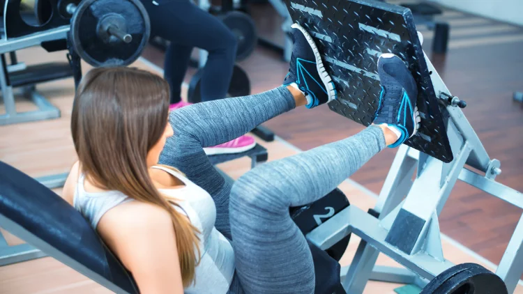 A woman is wearing a gray top, gray leggings, and blue shoes while performing the wide leg press foot placement exercise, she is sitting on a leg press machine with her legs spread wide apart, and her toes pointing outward.
