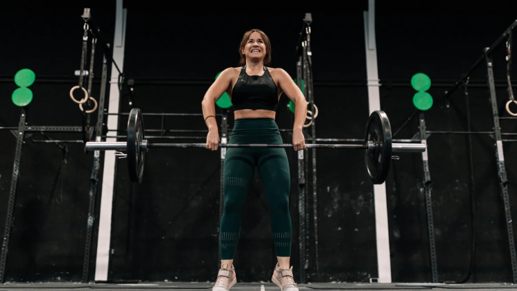 A woman wearing a black top, green pants is performing the hang clean in fifth position, with her arms lowering the barbell to the ground.