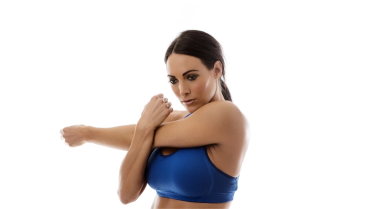 A woman wearing a blue sports bra stretches one arm with the help of her other arm in white background.