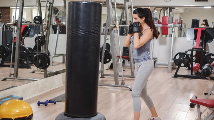 A young girl with her hair pulled back in a ponytail is standing in front of a black punching bag with her fists wrapped in black training gloves, she wears a gray gym top and matching leggings, while other gym equipment, such as weights and machines, are visible in the background.