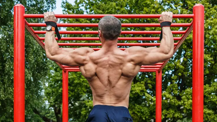 A topless man wearing blue pants performing a wide grip pull ups exercise on a bar structure at the park.