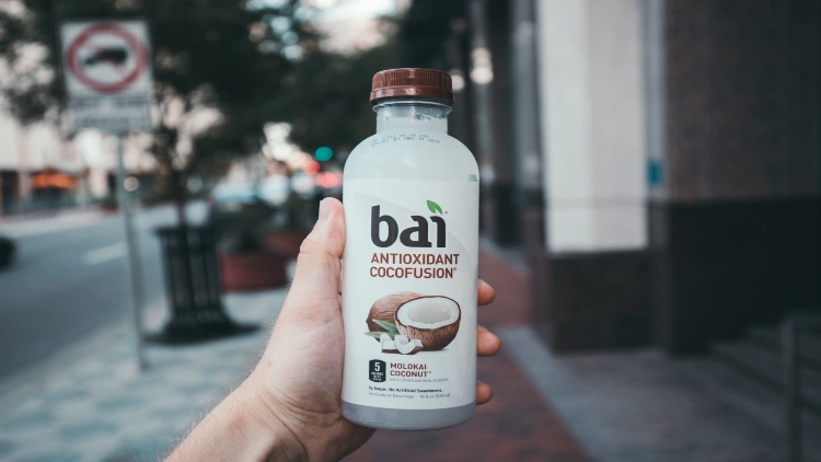 A hand holding a bottle of bai drink with a blurred street background, the bottle has a white label and brown cap, and the drink inside is a vibrant white color.