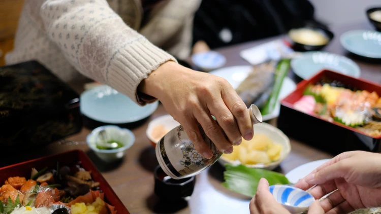 A hand is carefully and slowly pouring sake from a traditional ceramic sake vessel into a small ceramic sake cup with intricate designs and patterns, suggesting an authentic Japanese aesthetic; the background of the image is blurred, hinting at a meal or social gathering, suggesting that the sake is being enjoyed.