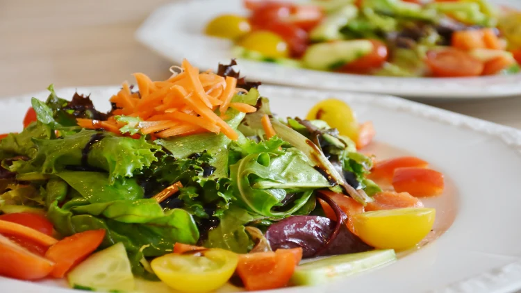 Two servings of a healthy vegetable salad made with green leafy vegetables, sliced tomatoes, and finely chopped carrots served on white plates.