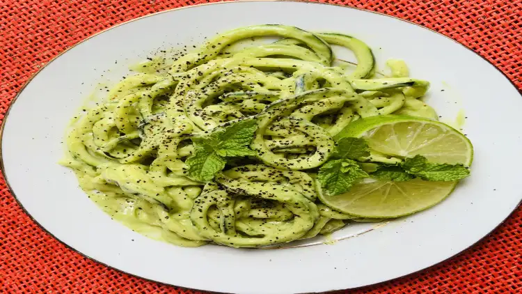 A white plate serves zucchini noodles in a creamy sauce, topped with fresh parsley leaves, ground black pepper, and two sliced green lemons on the side, and is placed on a red tablecloth in the background.
