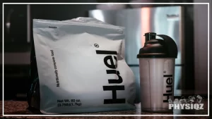 A bag of Huel, a powdered meal replacement, and a Huel shaker bottle, both placed on a white countertop in a kitchen, the bag features the Huel logo and nutritional information, while the shaker bottle has a black lid and clear body with measurements marked on it, the blurred background shows various kitchen appliances.