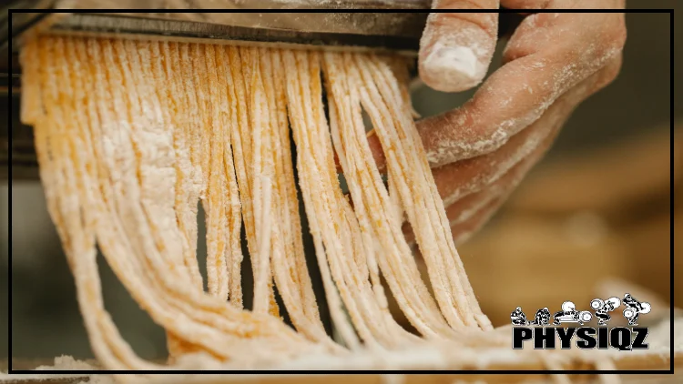 A white man's hand has long nails, is covered in dusty white flour and is holding a pasta maker with yellow tinted noodles stringing out from the silver blades which makes him wonder if chickpea pasta is keto or not.