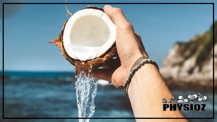 A white person holding half of a freshly opened coconut and considers is coconut water ketogenic, as he pours out its clear water into the blue seas on a tropical island with green forage on the rocks.