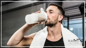 Muscular man with short hair wearing a black tank top and a white towel around his neck is depicted holding a white protein shake tumbler in his right hand and drinking from it, the shake appears to have a thick and creamy consistency, and is likely packed with protein and nutrients for post-workout recovery, the background is blurry, suggesting that the man is in a gym or fitness center.