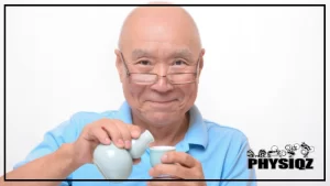 An elderly Japanese man with a bald head is wearing a light blue shirt and eyeglasses as he smiles at the camera, in his hand, he holds a traditional Japanese sake cup while preparing to pour from a vessel beside him, his smile is warm and welcoming, with crinkles forming around his eyes.