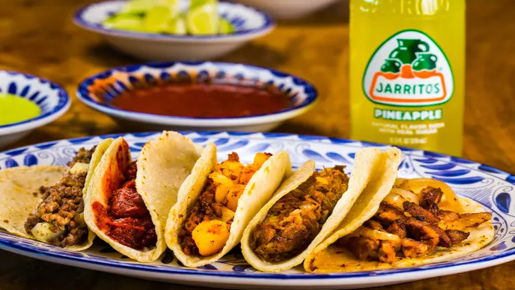 Freshly baked tacos with beef, onion, cheese, and meat are served on a plate with blue and white feather-like patterns on the edges, with a bottle of Jarritos' pineapple, a plate of ketchup, cut lemons and mustard in the background, placed on a wooden tabletop.