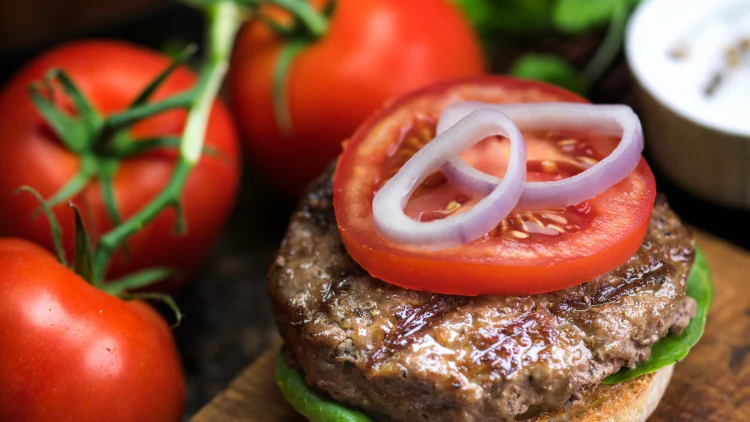 Juicy grilled beef patty topped with a slice of tomato and two slices of onion, the beef patty is thick and has visible grill marks on its surface, the tomato slice on top of the patty is red and ripe, while the onion slices are white and have a slightly translucent appearance, in the background, three more tomatoes can be seen, adding to the freshness, the background is slightly blurred, keeping the focus on the beef patty and its toppings.