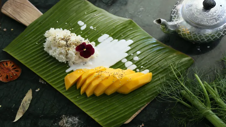 A dish consisting of seven slices of mango, sticky rice, and coconut milk served on a banana leaf and displayed on top of a dark shiny surface with a silver kettle and a green leafy vegetable on the side.