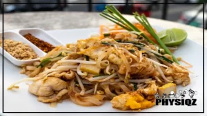 A serving of low carb thai food such as the Pad Thai dish consisting of noodles, peanuts, tamarind paste, and a variety of other ingredients including chicken, tofu, and lime, presented on a white plate and served on a table outside in the fresh air.