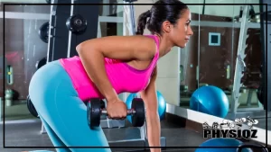 A woman with braided hair, wearing a pink tank top and blue pants is performing one arm dumbbell bent over row workout, one of lower back exercises with dumbbells, at a gym with a mirror in front of her.