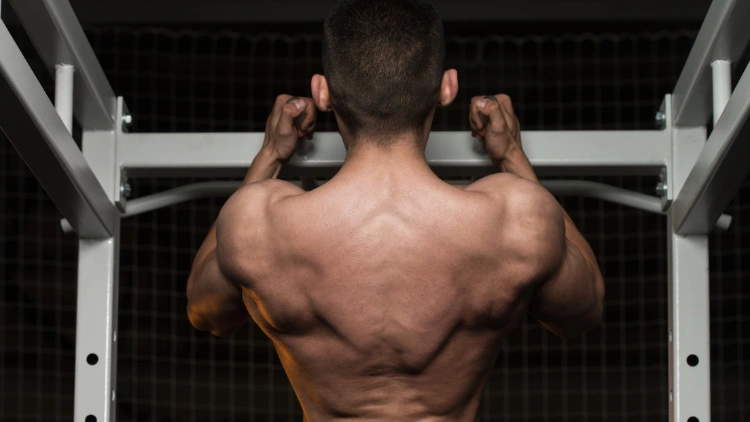 A back view of a topless man doing pull ups exercise using a neutral grip in a dimly lit gym.