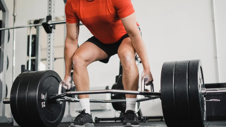 A man wearing a red activewear shirt, black short and shoes with white socks is performing a deadlift using a trap bar with three weighted plates in a gym with white wall.