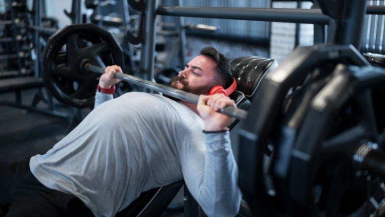 A bearded man in gray sweatshirt wearing a red headphones in his head doing a close grip bench press, various gym equipment are seen in the background.